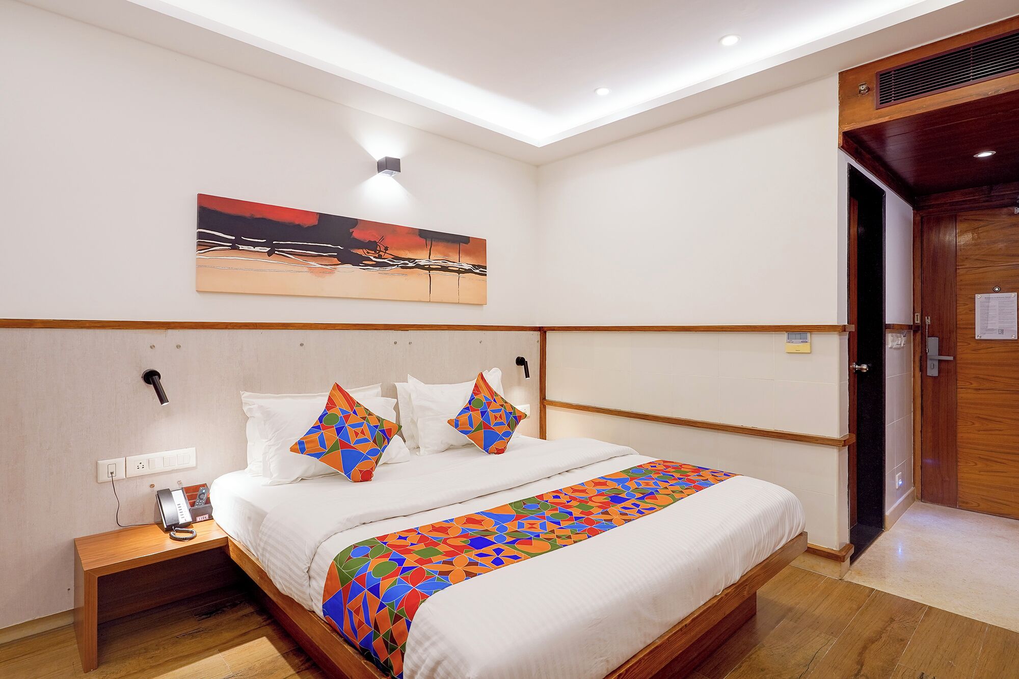 A hotel room with a bed, a bedside table with a phone, a painting on the wall, and a wooden door.