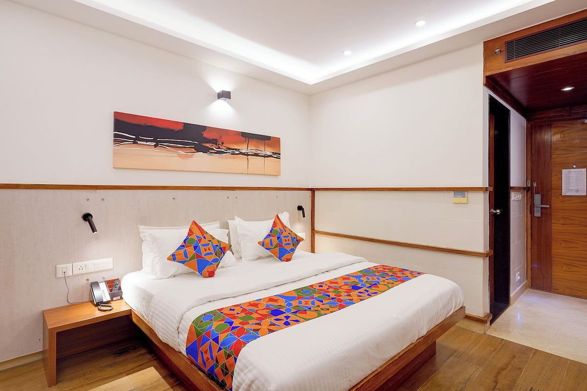 A hotel room with a bed, a bedside table with a phone, a painting on the wall, and a wooden door.