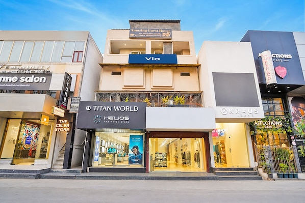 A multi-story building with various storefronts including a watch store, a cafe, and a clothing store. - Via Amazing Guest House (New Delhi)