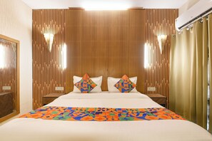 A hotel room with a wooden panel wall, a bed with a colorful geometric-patterned bedspread, two pillows with geometric designs, a nightstand with a lamp, and a window with curtains. - Via Gem 92 (New Delhi)
