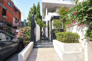Estret camí urbà que condueix a un edifici de façana blanca, envoltat de vegetació i flors roses.