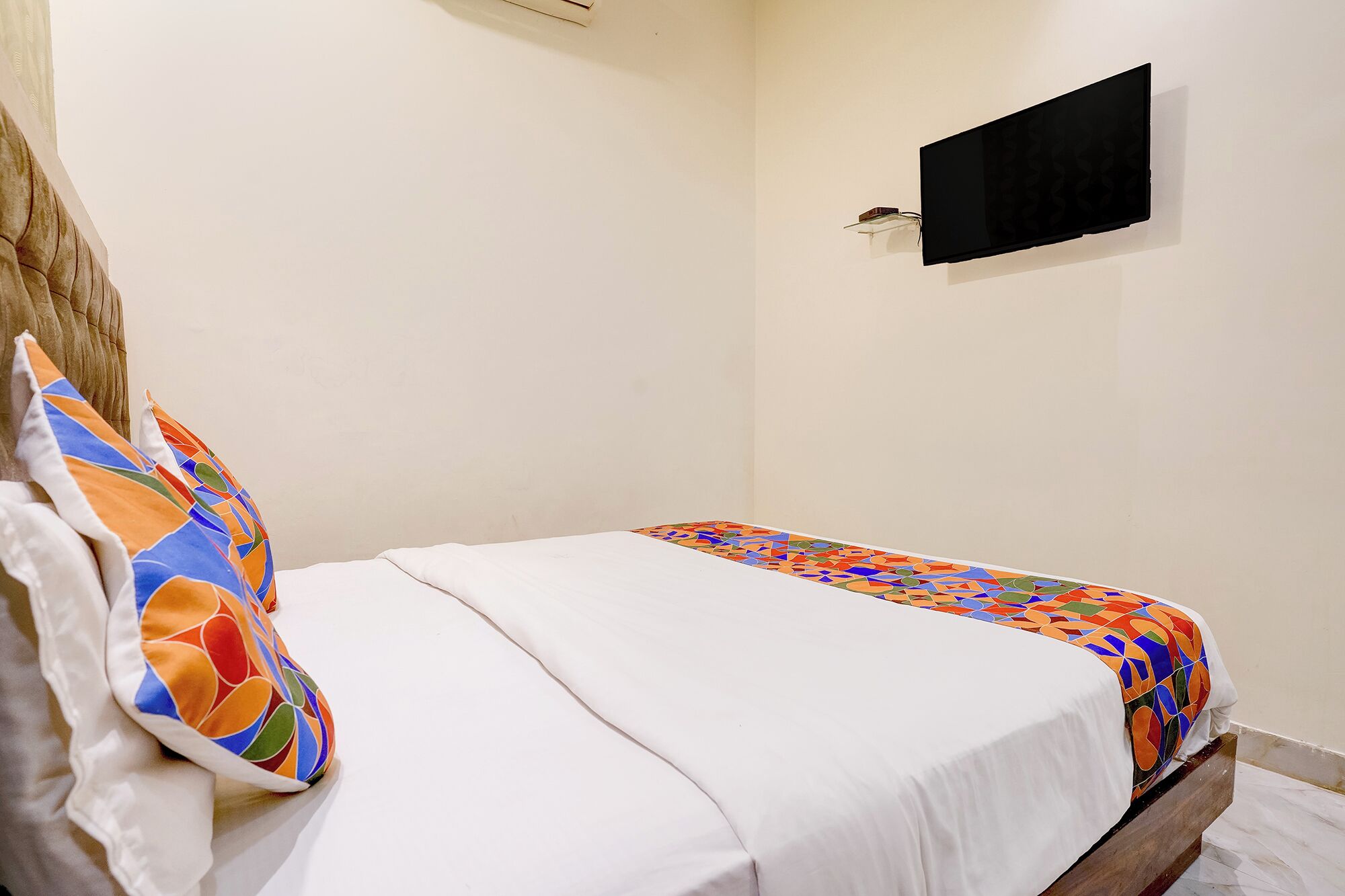 A single bed with a colorful bedspread, a white pillow, and a plain wall with a flat-screen TV mounted above it.