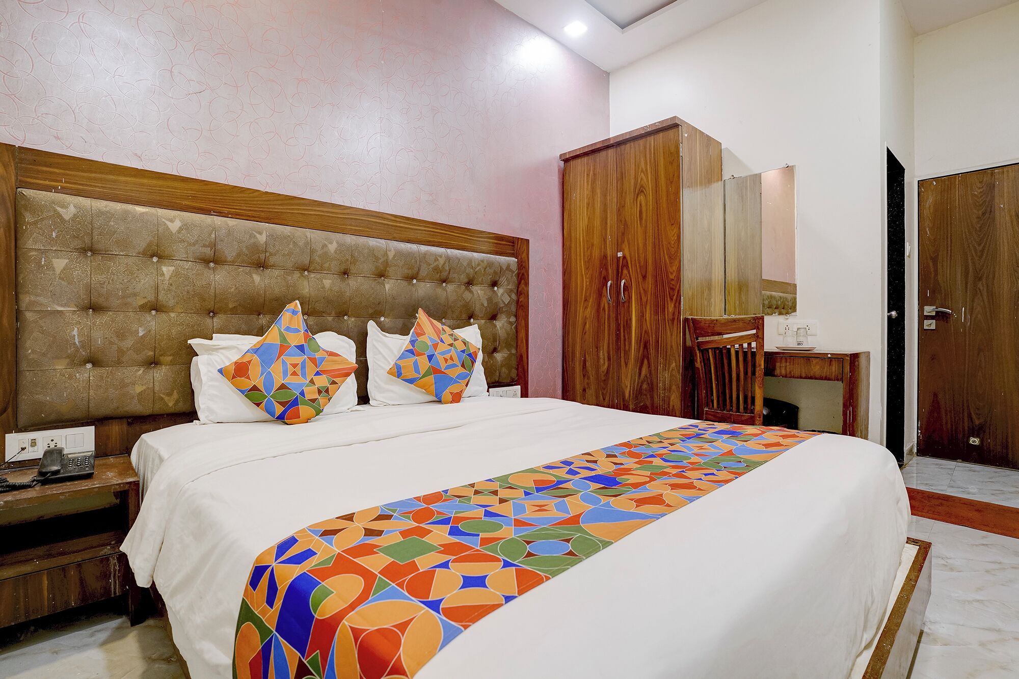 Une chambre à coucher avec une tête de lit capitonnée, un lit agrémenté d’oreillers à motifs, une armoire en bois et un bureau avec une chaise.