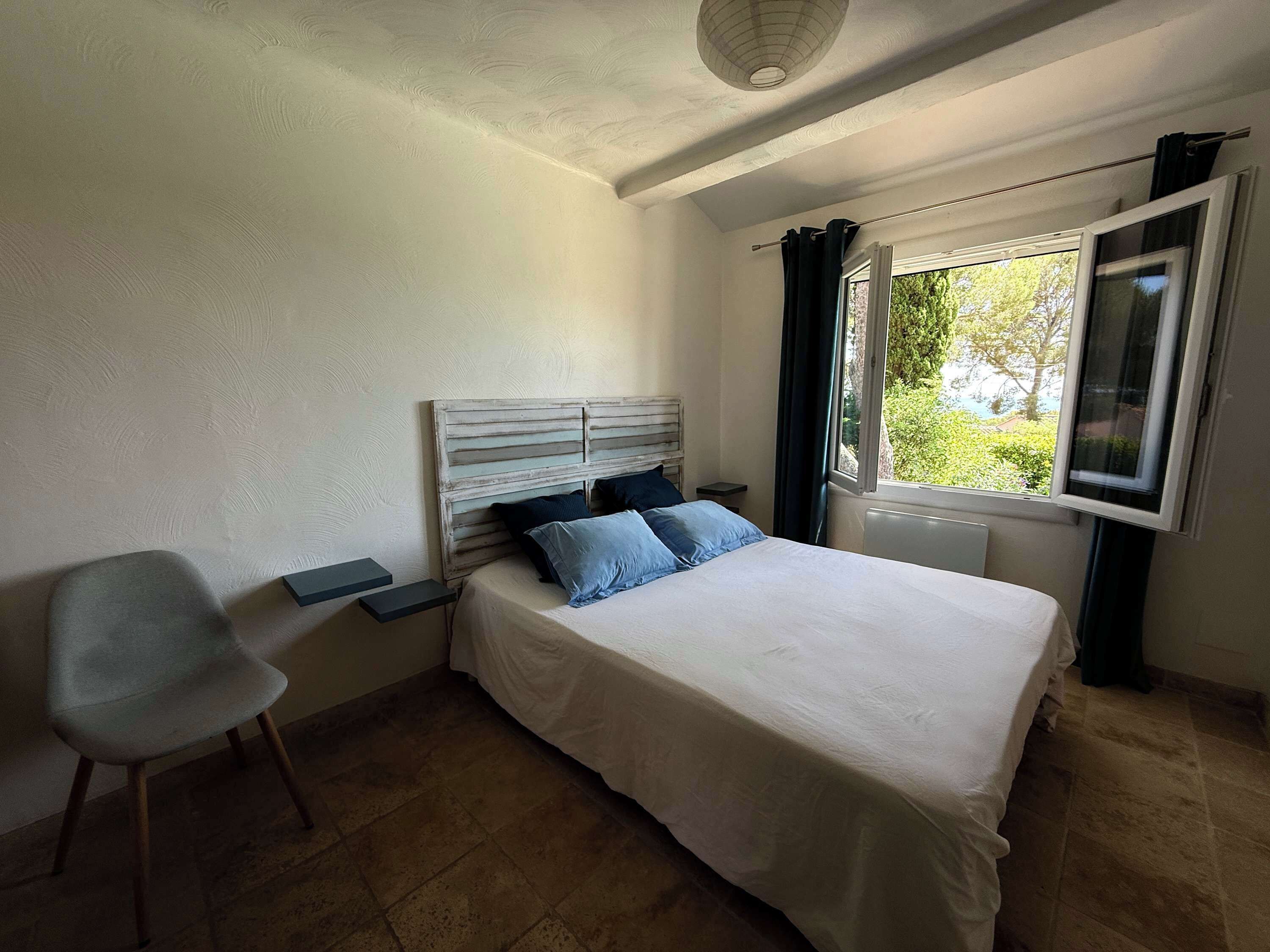 Een slaapkamer met een bed, een stoel, een nachtkastje, een raam met gordijnen en een plafondlamp.