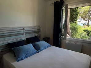 Ein Schlafzimmer mit Bett, Nachttisch, einem Fenster mit Vorhängen und Blick auf Grünfläche.