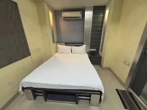 Ein schmales Hotelzimmer mit einem Einzelbett, einer Klimaanlage, einem Nachttisch und einem kleinen Regal.