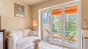 A room with a sofa, a small round table, a wall-mounted light, a sliding glass door to an outdoor area with a table and chairs, and a framed picture on the wall. - Newly studio apartment in a quiet residence (Mandelieu-la-Napoule)