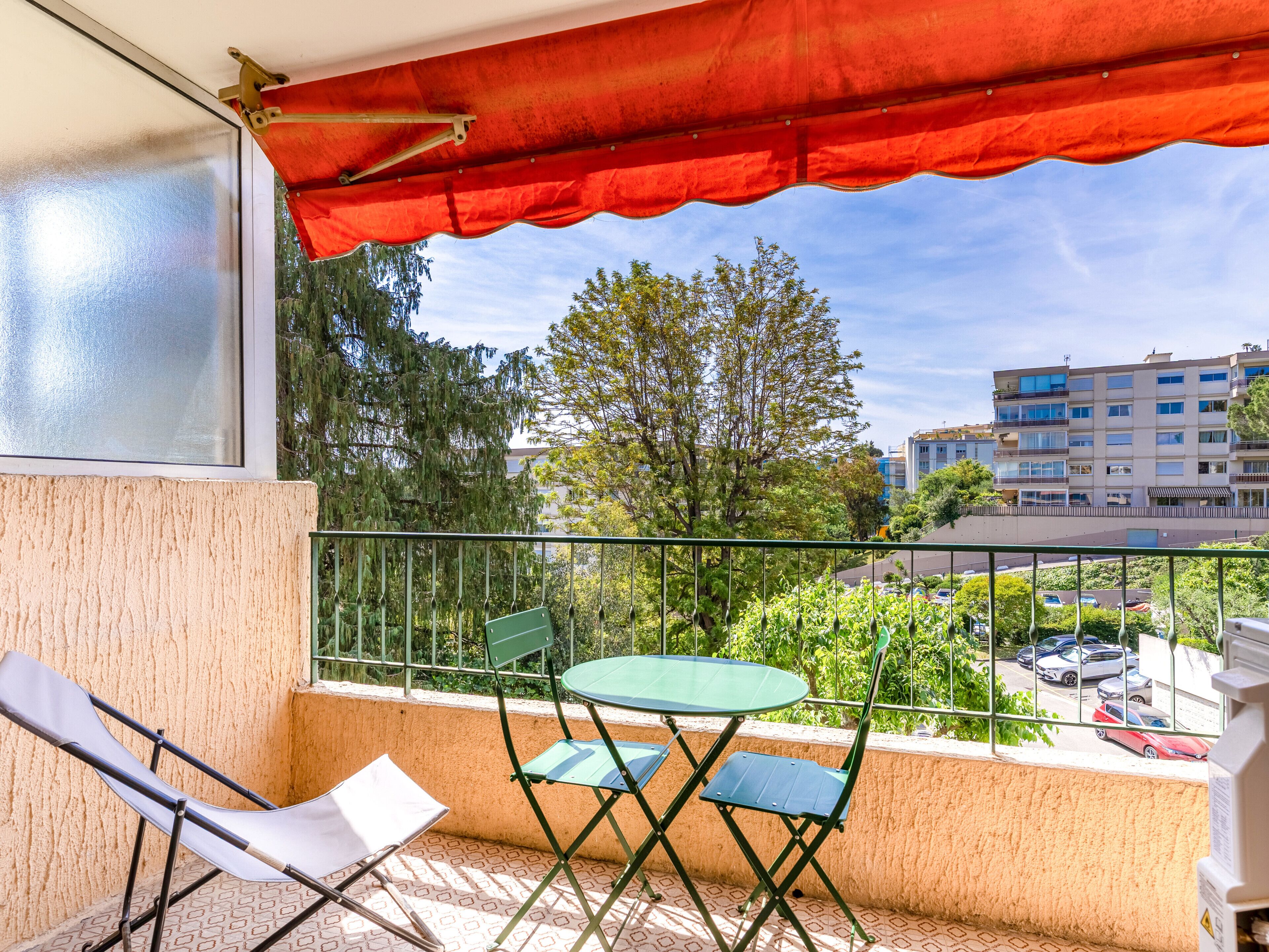 Un balcon doté d’un auvent rouge, de deux chaises vertes, d’une table blanche et offrant une vue sur un paysage urbain avec des bâtiments et des arbres.
