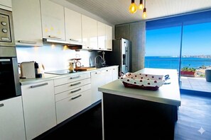 Eine moderne Küche mit weißen Schränken, einem Edelstahl-Kühlschrank und Blick aufs Meer.