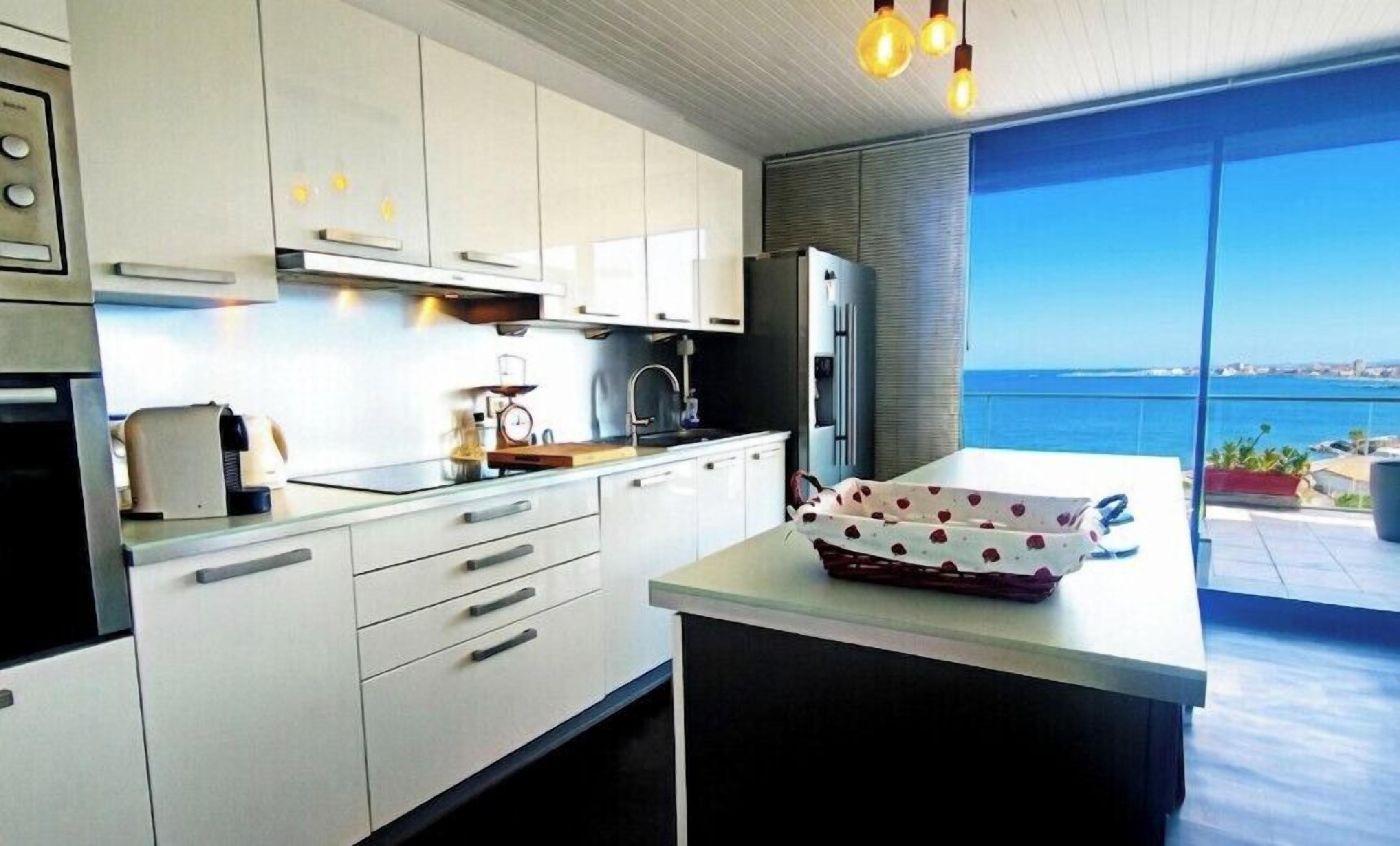 A modern kitchen with white cabinets, a large island, and a view of the sea.