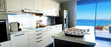A modern kitchen with white cabinets, a large island, and a view of the sea.