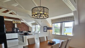 A dining area with a wooden table and chairs, a chandelier, and a kitchen in the background.