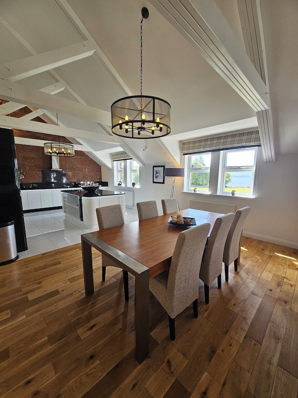 A dining area with a wooden table and chairs, a chandelier, and a kitchen in the background.