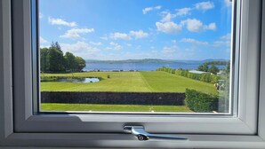A window with a view of a green landscape, a clear sky, and a distant body of water.