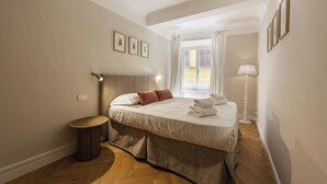 Ein Schlafzimmer mit Bett, Nachttischlampe, einem Fenster mit Vorhängen und einem kleinen runden Tisch.