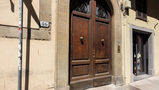 Eine doppelte Holztür mit einem Schild mit der Aufschrift '46' darüber, eine Metallstange mit Aufklebern und ein kleines Fenster mit schmiedeeisernem Zierwerk.