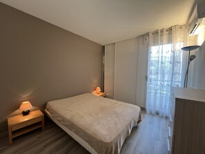 A bedroom with a bed, bedside lamps, a wooden nightstand, a window with sheer curtains, and a ceiling-mounted air conditioner. - Cesaree Residence – Renovated 2-room apartment with pool view, air conditioning, garage and terrace – Sleeps 4. (Fréjus)