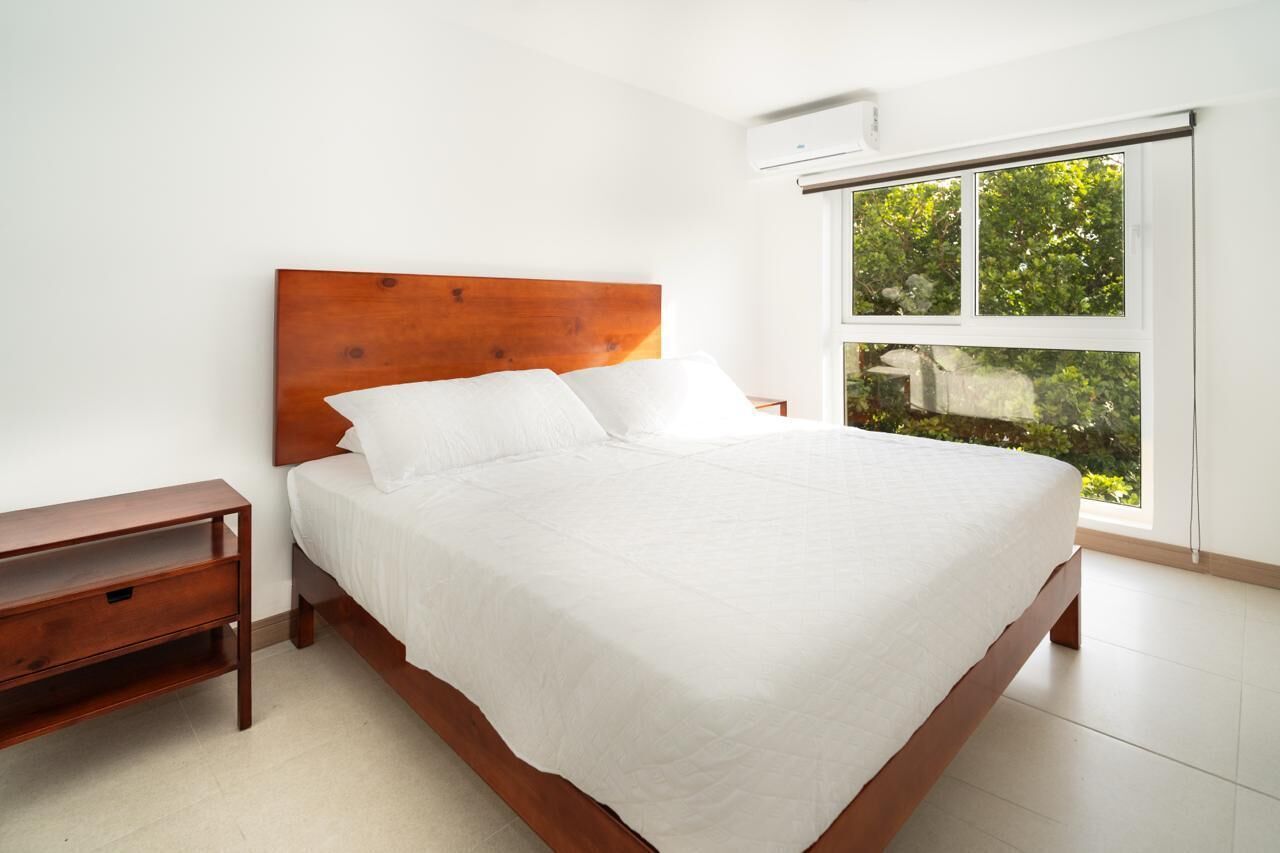 Une chambre à coucher avec une tête de lit en bois, un couvre-lit blanc, une table de chevet et une fenêtre donnant sur un paysage verdoyant.