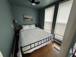 Quarto com cama, mesa de cabeceira, abajur, ventilador de teto e janela com persianas.