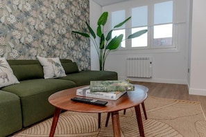 A living room with a sofa, a coffee table with books and a remote, a large plant, and a radiator.