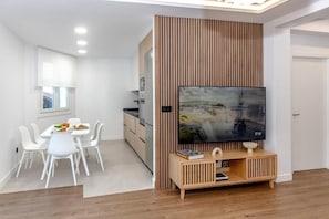 A modern living room with a flat-screen TV, wooden panel wall, white dining set, and a wooden console table.