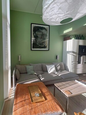 A living room with a sofa, a coffee table, a painting on the wall, and a kitchen area in the background.