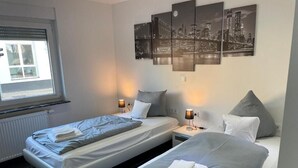 Une chambre d’hôtel avec deux lits, une fenêtre avec des stores, une œuvre d’art représentant un paysage urbain accrochée au mur et un radiateur.