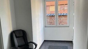 Une pièce avec un fauteuil de bureau noir, une grande fenêtre donnant sur un immeuble en briques et un tapis foncé sur le sol.