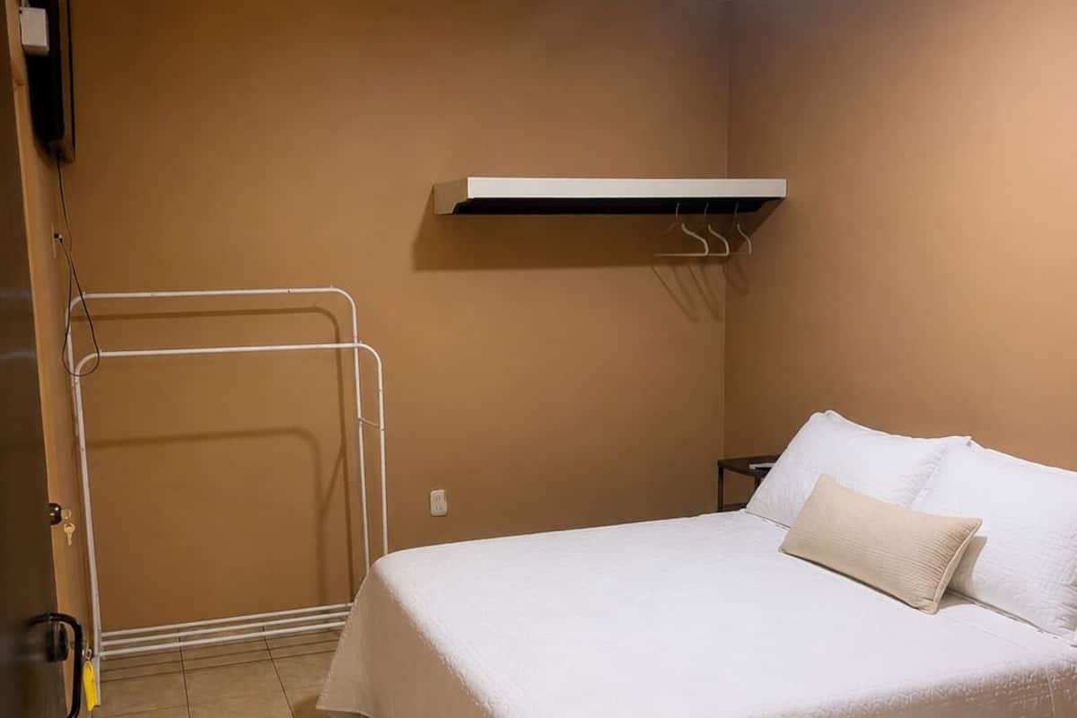 Habitación de hotel con cama, perchero, calefactor y unidad de aire acondicionado instalada en la pared.