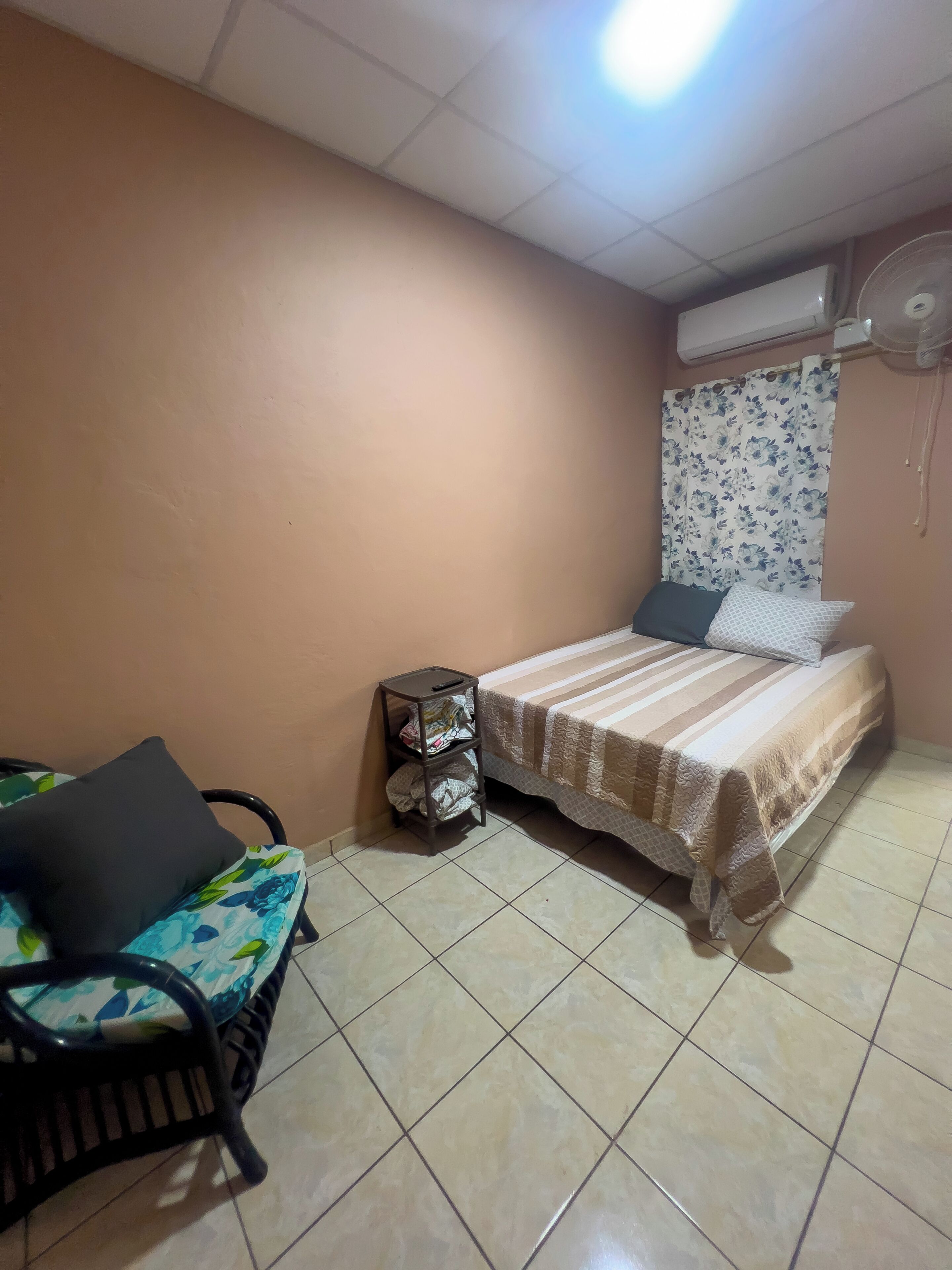 A bedroom with a bed, a chair, a small table, and an air conditioning unit.