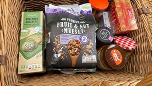 A basket containing various food items including a pack of Rivercote Rosemary Crackers, a bag of Premium Fruit & Nut Muesli, a jar of Delgarde Raspberry Presage, and a box of Roobos. - Cosy Caravan Sleeps 2 With Patio and Parking (Kilteean)
