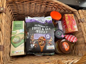 Una cesta con varios alimentos, incluyendo una caja de galletas Rivercote Rosemary, una bolsa de avena Premium con frutas y nueces, un tarro de mermelada de frambuesa Delgarde y una caja de té Roobos.