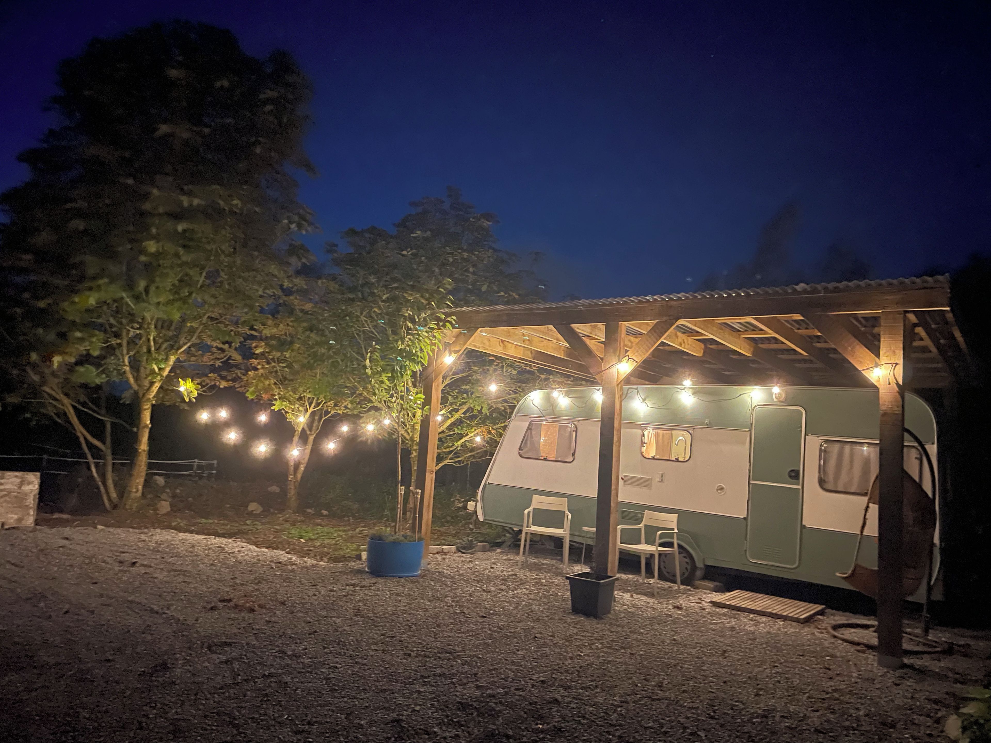 A covered RV with string lights, a gravel ground, and trees in the background.
