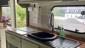 Una kitchenette compacta con un lavamanos, un escurreplatos, un cubo de basura pequeño y una ventana con vistas a una casa y follaje.