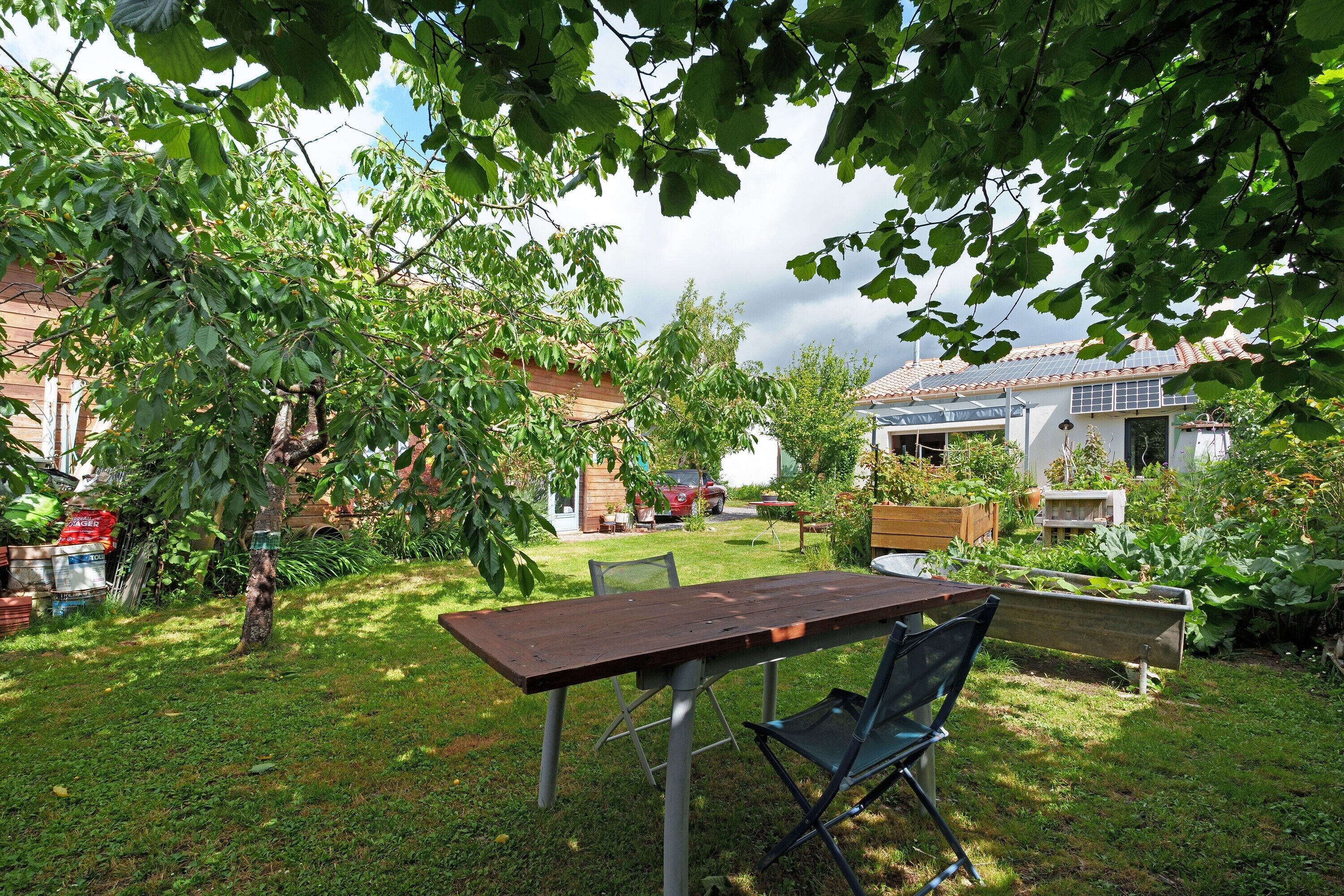 Eine Terrasse mit Tisch und Stühlen, umgeben von viel Grün und einem Haus im Hintergrund.