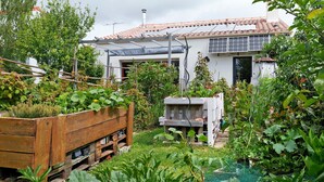 Un jardín con una jardinera de madera, un edificio blanco con tejado de tejas y una estructura de madera al fondo.