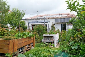 A garden with a wooden planter, a white building with a tiled roof, and a wooden structure in the background.