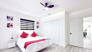 Un dormitorio moderno con una cama, paredes blancas, un ventilador de techo y una puerta que conduce a otra habitación.