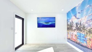Una habitación con piso de madera, un televisor grande montado en la pared, y un mural colorido en la pared contigua.