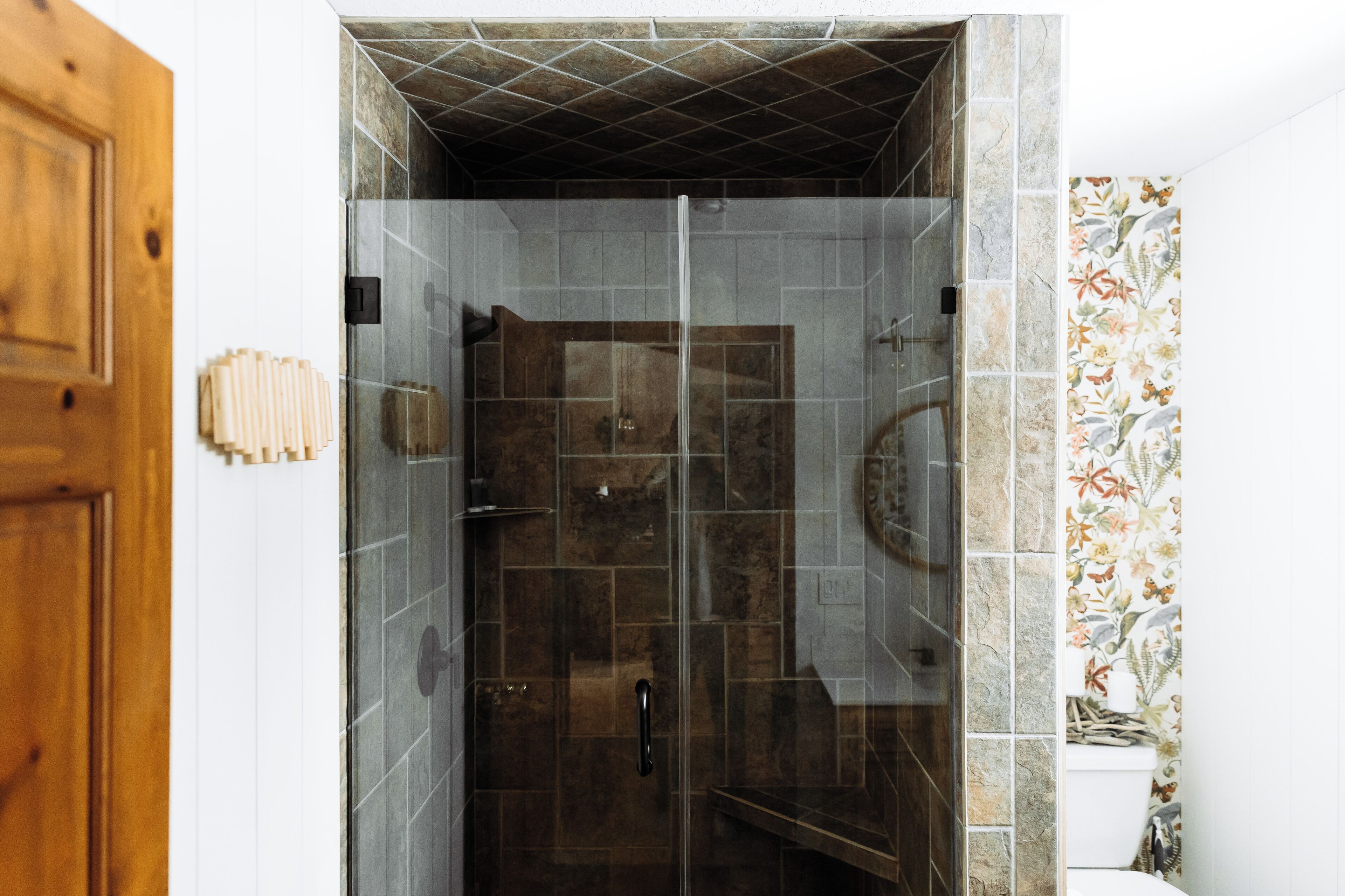 Une douche avec des portes en verre, une porte en bois et un mur carrelé orné d’un motif floral.