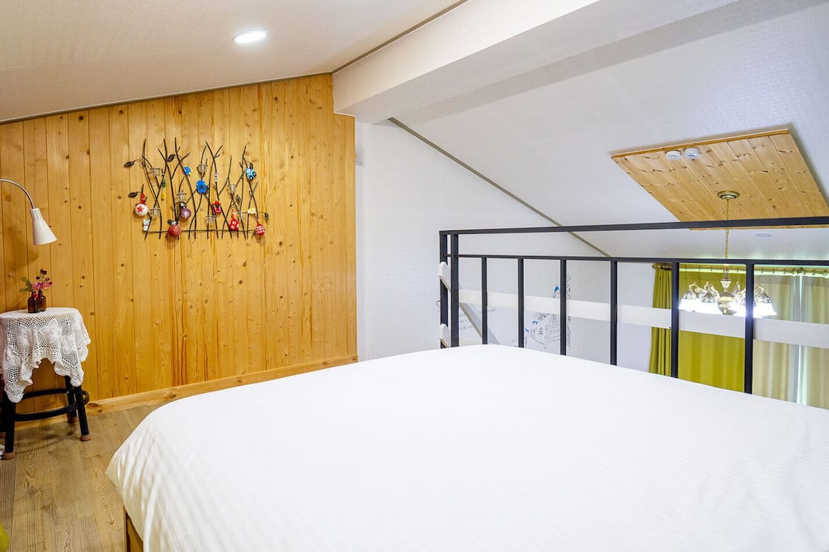 A bedroom with a wooden wall, a bed, a small table with a lamp, and a balcony with a view.