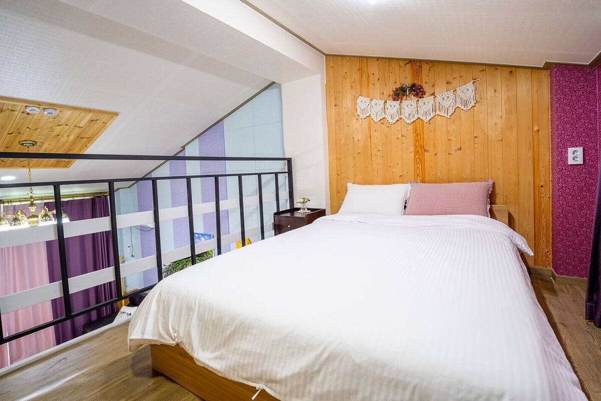 A bedroom with a wooden headboard, a bed with white bedding, a small nightstand, and a balcony with purple curtains.