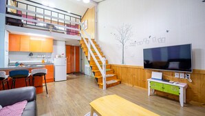 A spacious living area with a wooden staircase, a kitchen with orange cabinets, a dining area with bar stools, a flat-screen TV, and a small table with a calendar. - Supia Pension (Pyeongchang)