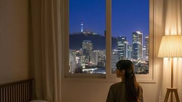 A room with a bed, a desk, a lamp, and a window with a city view.