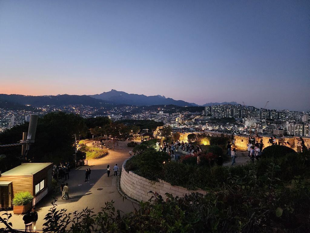 A cityscape at dusk with a historic site in the foreground featuring a paved walkway and a stone wall.