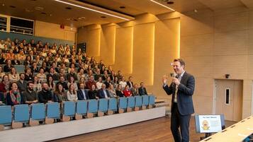Une salle de conférence avec des rangées de chaises bleues, un pupitre et un panneau avec du texte.