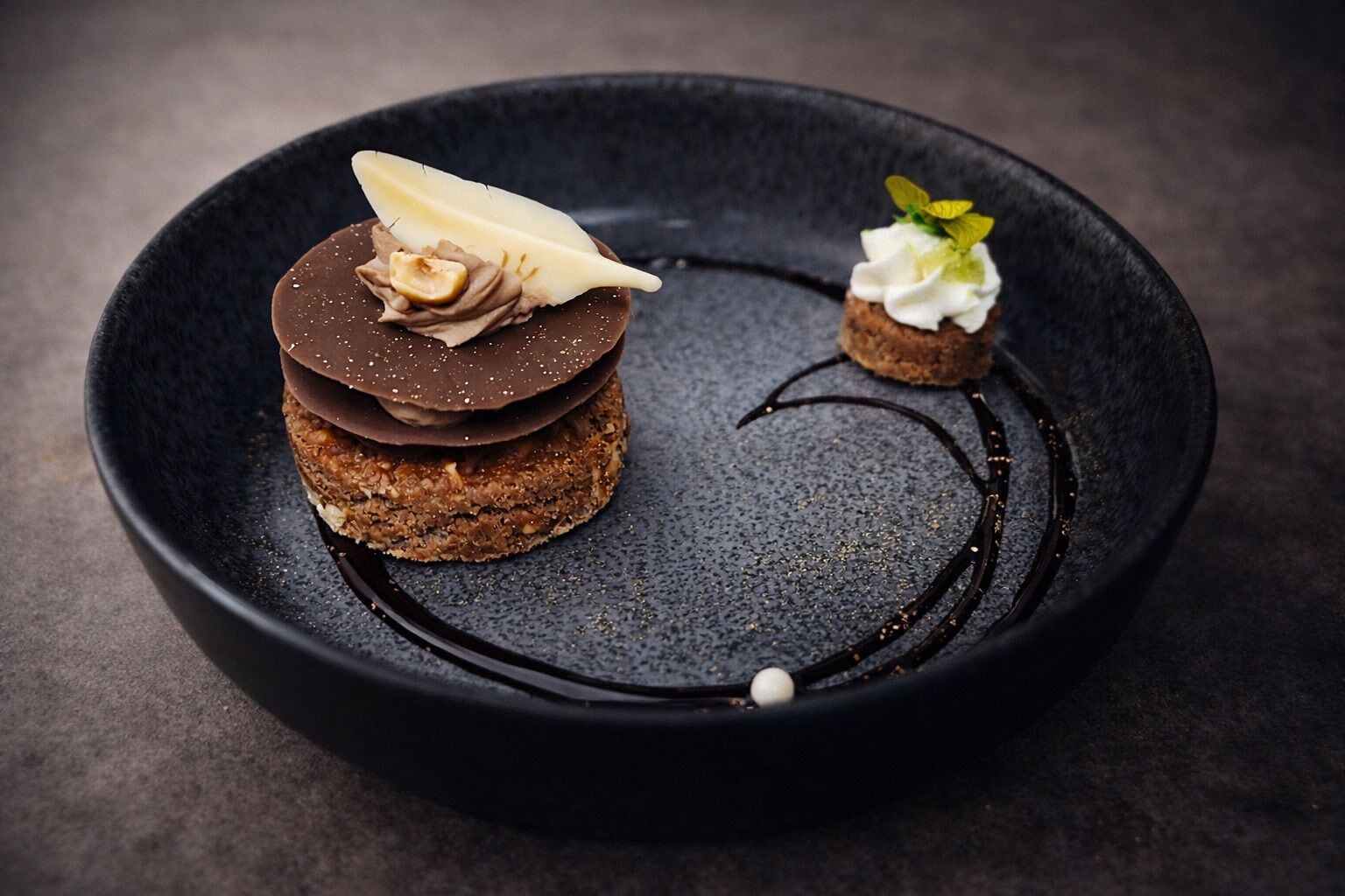 En dessert på en mørk tallerken med en chokoladebund, et bananskive og en lille klat fløde.