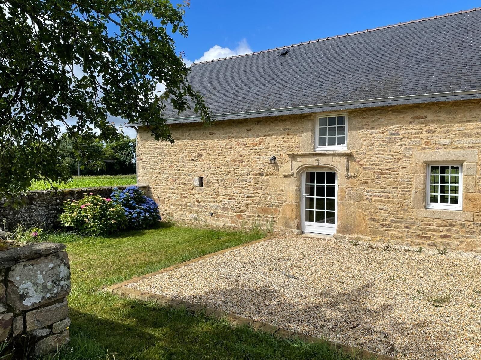 A stone building with a gravel driveway, a small garden with flowers, and a clear blue sky.