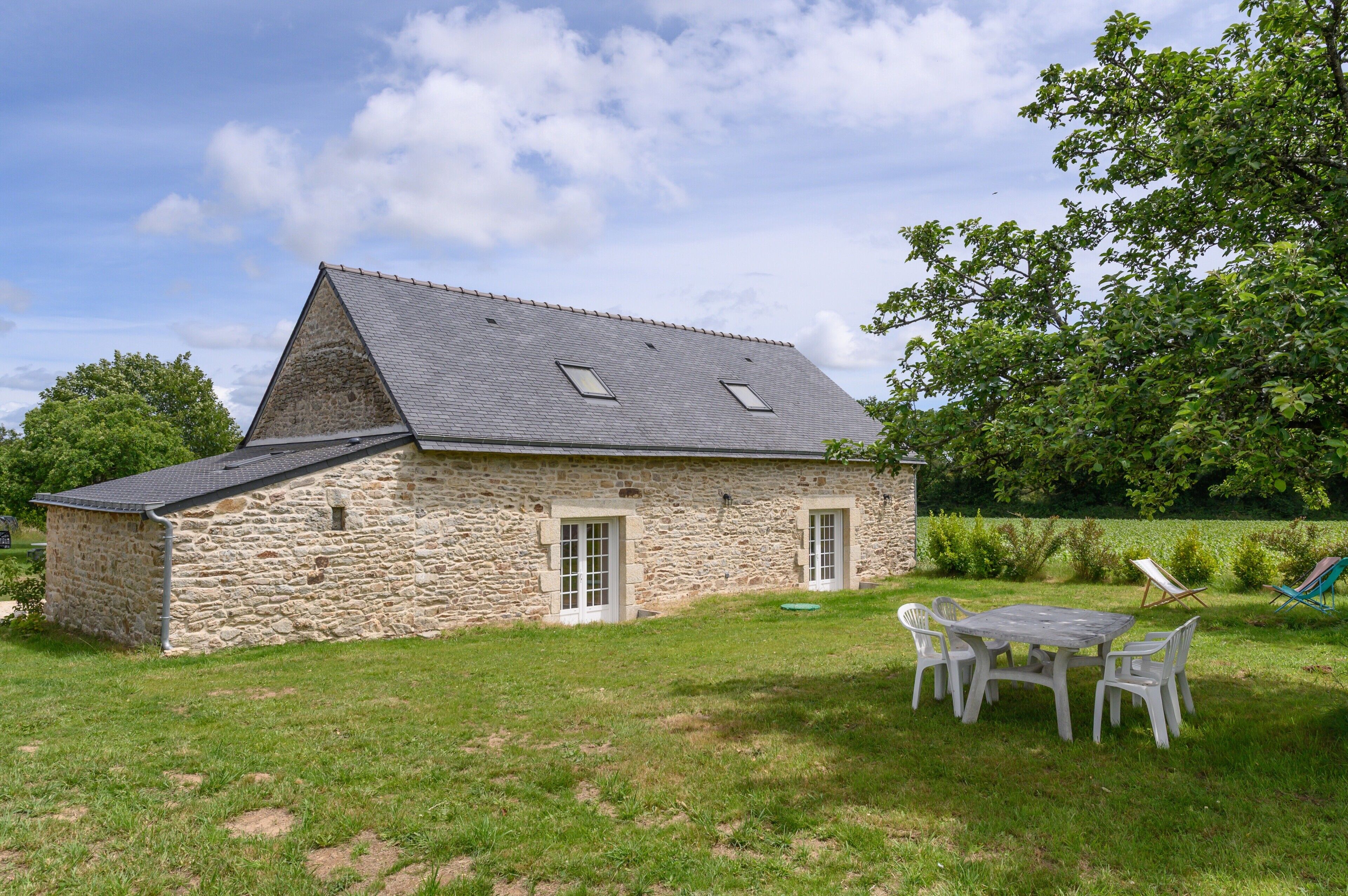 Een stenen huis met schuin dak, een kleine deur en een raam. Er is een gazon met een tafel en stoelen.