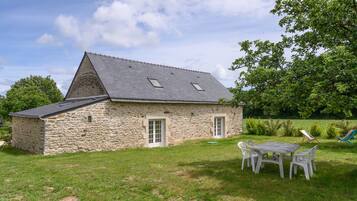 Een stenen huis met schuin dak, een kleine deur en een raam. Er is een gazon met een tafel en stoelen.